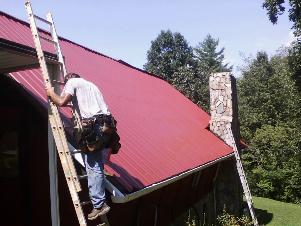 Roofer on a ladder beside a red metal roof and stone chimney — Soffit & Fascia Repair by Roofing Brothers in Wye Mills